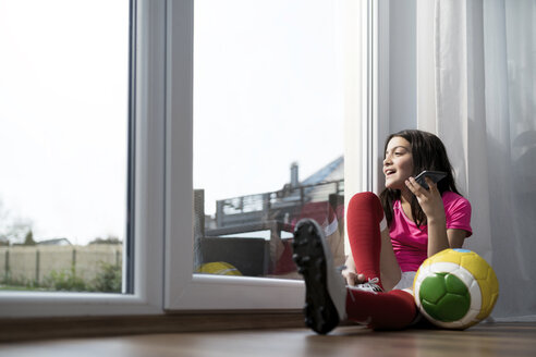 Mädchen im Fußball-Outfit sitzt auf dem Boden im Wohnzimmer und spricht mit jemandem auf ihrem Smartphone - SBOF00654