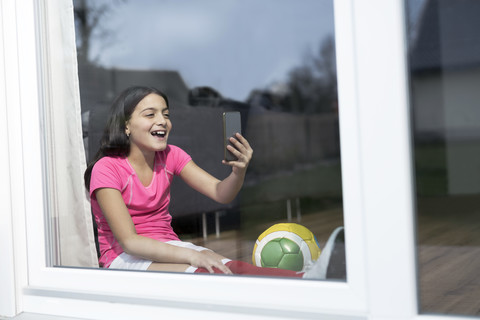 Glückliches Mädchen im Fußball-Outfit sitzt auf dem Boden im Wohnzimmer und macht ein Selfie, lizenzfreies Stockfoto
