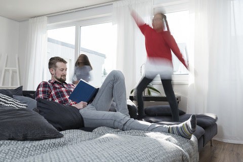 Mädchen springen auf dem Sofa im Wohnzimmer, während der Vater ein Buch liest, lizenzfreies Stockfoto