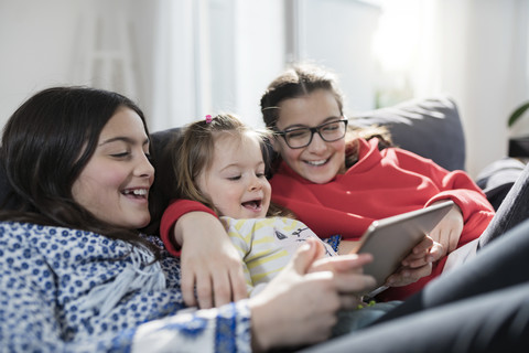 Drei Schwestern lächelnd und mit Tablet auf dem Sofa im Wohnzimmer, lizenzfreies Stockfoto