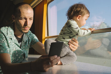 Father holding his baby girl and using smartphone while traveling by train - GEMF01815
