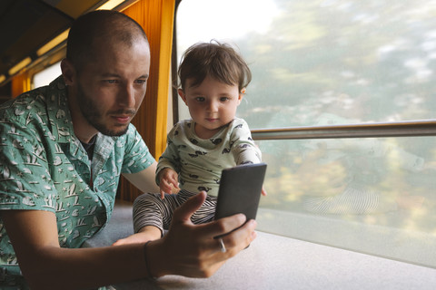 Vater hält sein kleines Mädchen und benutzt sein Smartphone während einer Zugfahrt, lizenzfreies Stockfoto