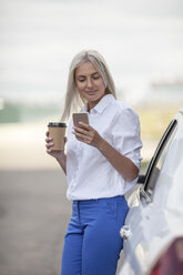 Smiling businesswoman with takeaway coffee and cell phone outside car - VPIF00053