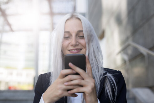 Lächelnde junge Geschäftsfrau, die in der Stadt ihr Mobiltelefon überprüft - GIOF03194