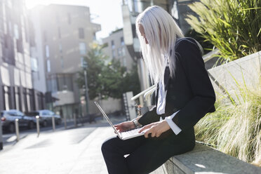 Junge Geschäftsfrau sitzt auf einer Mauer in der Stadt und benutzt einen Laptop - GIOF03204