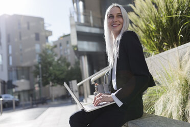 Lächelnde junge Geschäftsfrau, die auf einer Mauer in der Stadt sitzt und einen Laptop benutzt - GIOF03205