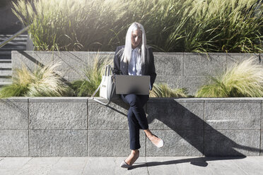 Young businesswoman sitting on a wall in the city using laptop - GIOF03206