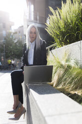 Junge Geschäftsfrau sitzt auf einer Mauer in der Stadt und benutzt einen Laptop - GIOF03207