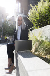 Young businesswoman sitting on a wall in the city using laptop - GIOF03207