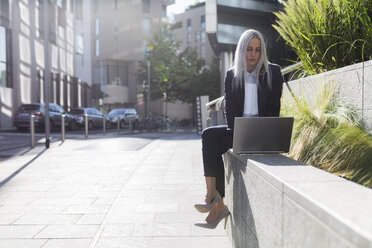 Junge Geschäftsfrau sitzt auf einer Mauer in der Stadt und benutzt einen Laptop - GIOF03208