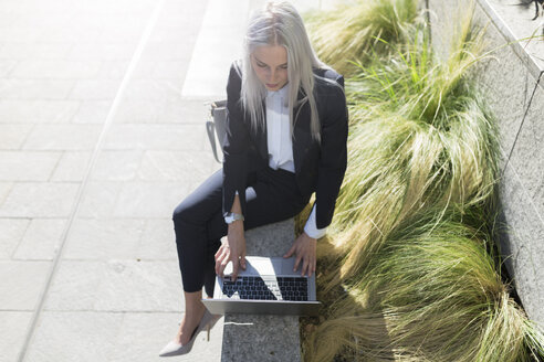 Junge Geschäftsfrau sitzt auf einer Mauer in der Stadt und benutzt einen Laptop - GIOF03209