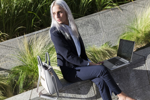 Young businesswoman sitting on a wall in the city using laptop stock photo