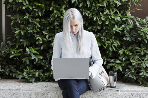 Junge Geschäftsfrau sitzt auf einer Mauer in der Stadt und benutzt einen Laptop - GIOF03224