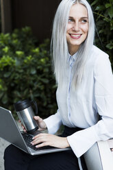 Smiling young businesswoman sitting on a wall with coffee mug and laptop - GIOF03225