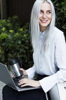Lächelnde junge Geschäftsfrau, die mit Kaffeetasse und Laptop an einer Wand sitzt - GIOF03225