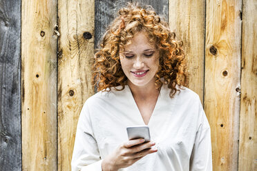 Porträt einer lächelnden jungen Frau, die vor einer Holzwand auf ihr Handy schaut - FMKF04474