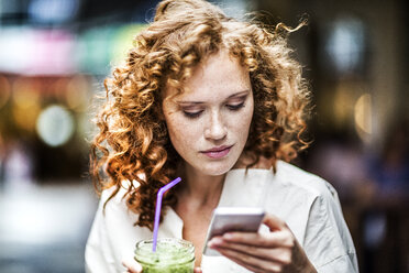 Porträt einer jungen Frau mit Getränk, die auf ihr Mobiltelefon schaut - FMKF04478