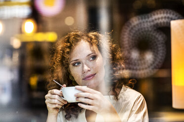 Porträt einer lächelnden jungen Frau mit Kaffeetasse hinter der Fensterscheibe eines Cafés - FMKF04482