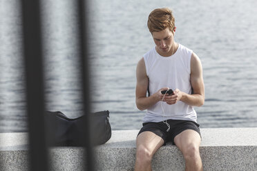 Junger Sportler sitzt mit Handy und Kopfhörern auf einer Mauer am Flussufer - VPIF00066