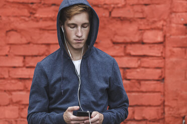 Young man with cell phone and earphones at red brick wall - VPIF00083