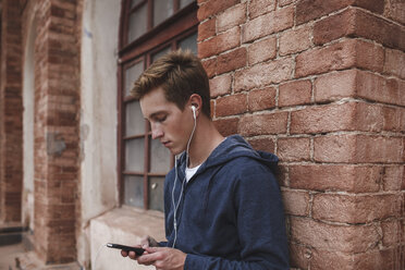 Junger Mann mit Mobiltelefon und Kopfhörern an einem Backsteinbau - VPIF00090