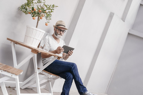 Älterer Mann mit Strohhut, der ein Tablet neben einem Tisch mit Orangenbaum benutzt, lizenzfreies Stockfoto