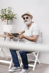 Mature man wearing straw hat holding tablet next to table with orange tree - JOSF01720