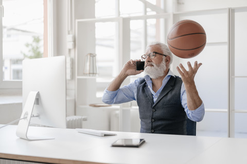 Älterer Mann am Handy am Schreibtisch spielt mit Basketball, lizenzfreies Stockfoto