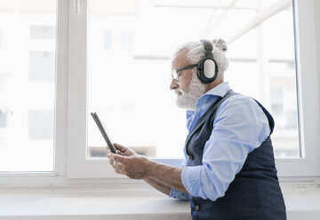 Älterer Mann mit Tablet und Kopfhörern am Fenster - JOSF01729