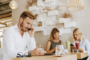 Mann am Mobiltelefon in einem Café mit zwei Frauen im Hintergrund - ZEDF00863