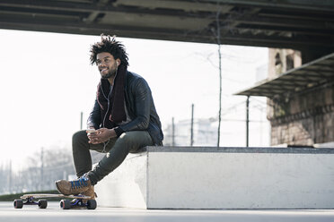 Smiling man with longboard sitting in skatepark under bridge holding a smartphone - SBOF00671