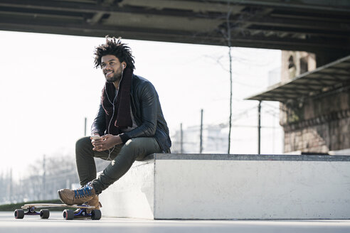 Lächelnder Mann mit Longboard sitzt im Skatepark unter einer Brücke und hält ein Smartphone - SBOF00671