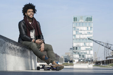 Lächelnder Mann mit Longboard, der im Skatepark sitzt und Musik auf seinem Smartphone hört - SBOF00678