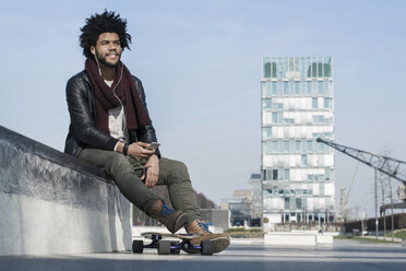 Smiling man with longboard sitting in skatepark listening to music on his smartphone - SBOF00678
