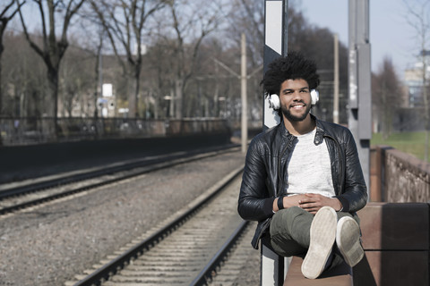 Lächelnder Mann, der an der Wand sitzt und neben den Eisenbahnschienen Musik hört, lizenzfreies Stockfoto