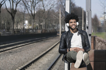 Smiling man sitting on wall listening to music next to train rails - SBOF00702