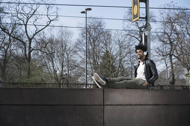 Smiling man sitting on wall listening to music next to train rails - SBOF00703