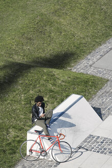 Mann sitzt im städtischen Skatepark mit Smartphone neben seinem Fahrrad - SBOF00709