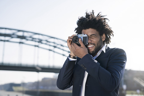 Lächelnder Mann im Anzug am Flussufer, der ein Foto mit einer alten Kamera macht, lizenzfreies Stockfoto