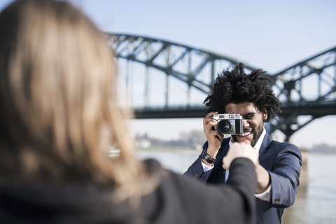 Lächelnder Mann im Anzug am Flussufer, der seine Freundin mit einer alten Kamera fotografiert, lizenzfreies Stockfoto