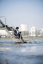 Geschäftsmann sitzt auf einer Treppe am Fluss und benutzt einen Laptop - SBOF00729