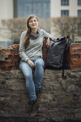 Portrait of smiling young woman sitting with her backpack on a wall - JSCF00006