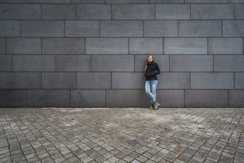 Junge Frau steht vor einer grauen Fassade und wartet, lizenzfreies Stockfoto