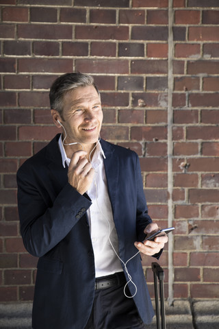 Älterer Geschäftsmann mit Kopfhörern und Smartphone an einer Backsteinmauer, lizenzfreies Stockfoto
