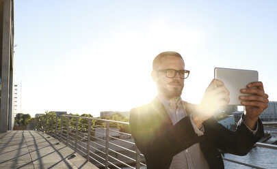 Businessman taking a selfie with tablet on a bridge in the city - FKF02548