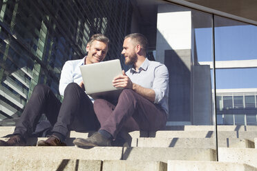 Two smiling businessmen sitting on stairs using laptop - FKF02587