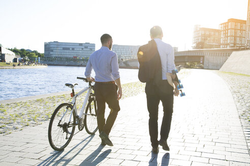 Two businessmen walking with bicycle and skateboard at the riverbank - FKF02590