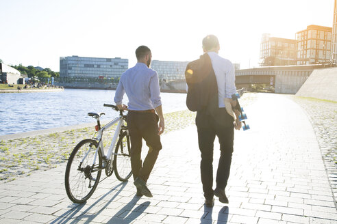 Zwei Geschäftsleute gehen mit Fahrrad und Skateboard am Flussufer spazieren - FKF02590