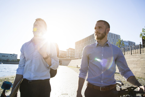 Two businessmen walking with bicycle and skateboard at the riverbank stock photo