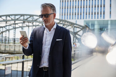 Deutschland, München, Porträt eines Geschäftsmannes mit Sonnenbrille, der auf sein Handy schaut - SUF00302